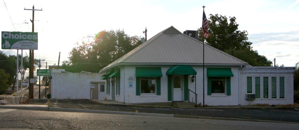 A small white building with green awnings sits beside a street, with an American flag and a Choices sign visible nearby.