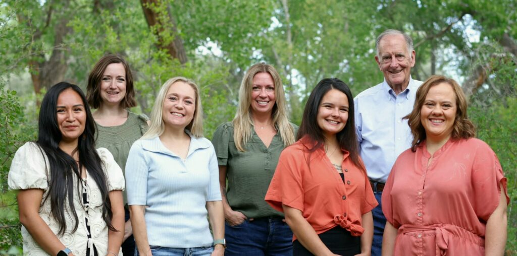 Seven adults stand together outdoors in front of green trees, with six women in the front and one man behind them.