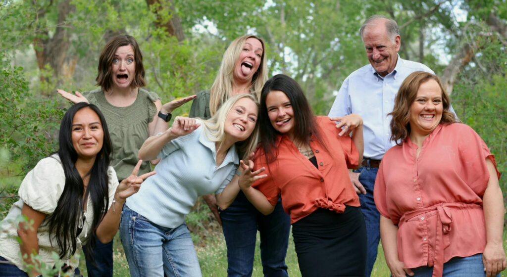 Seven adults pose together outdoors in a grassy park with trees in the background.