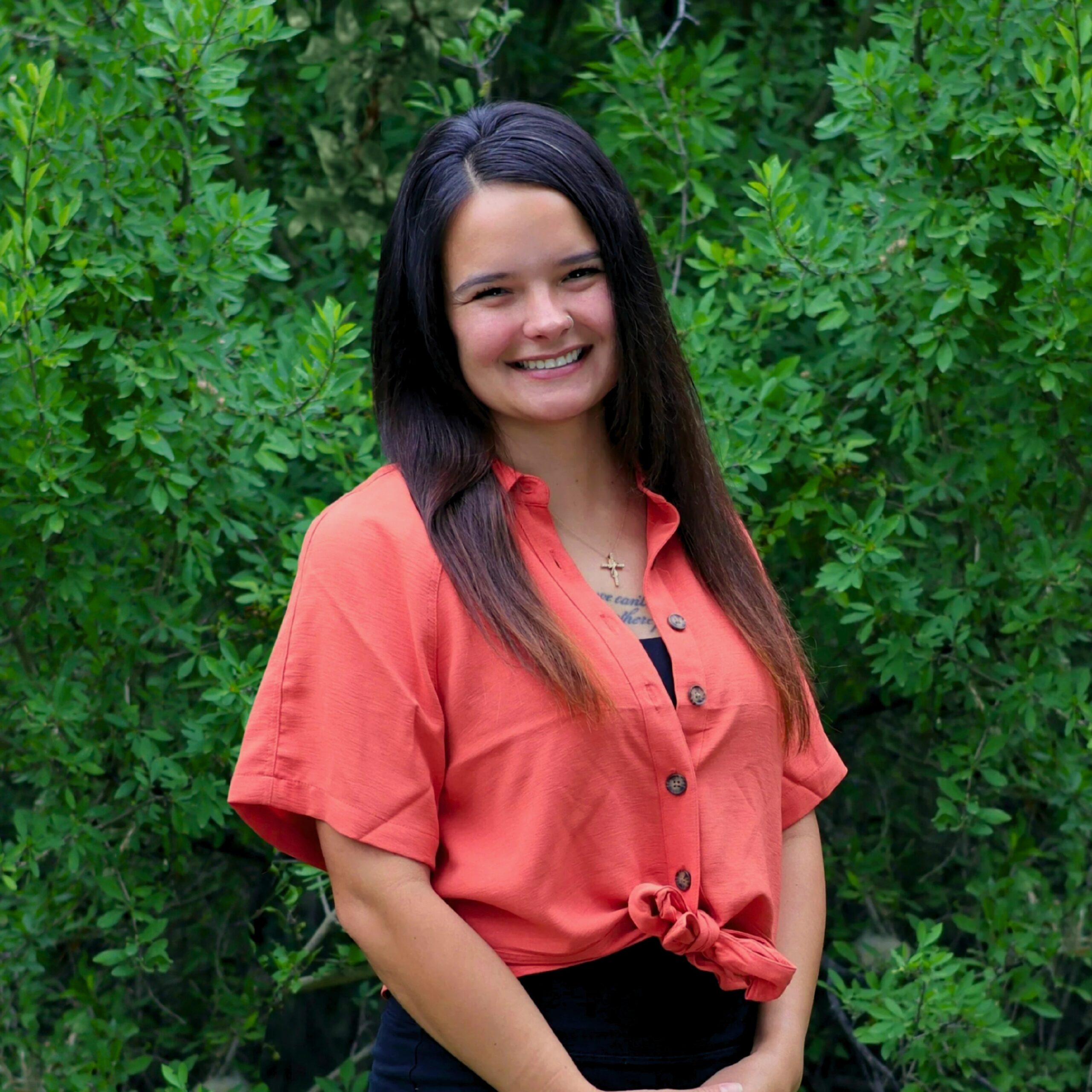 A smiling woman with long dark hair wears a coral button-up shirt tied at the waist, standing in front of dense green foliage.