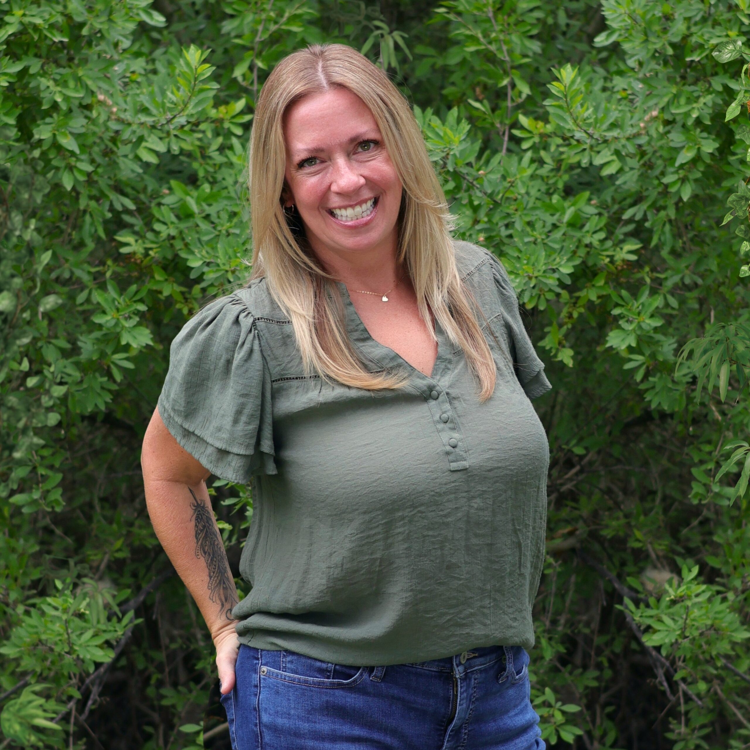 A blonde woman in a green blouse and jeans stands in front of dense green shrubs.