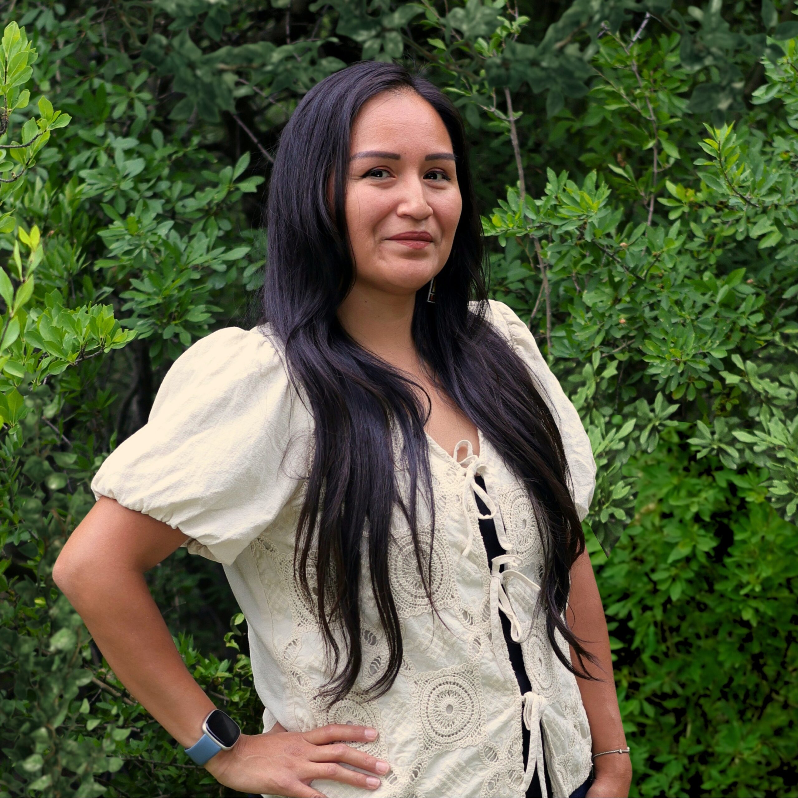 A woman with long dark hair stands outdoors in front of dense green foliage, wearing a cream blouse with puff sleeves and tying details.