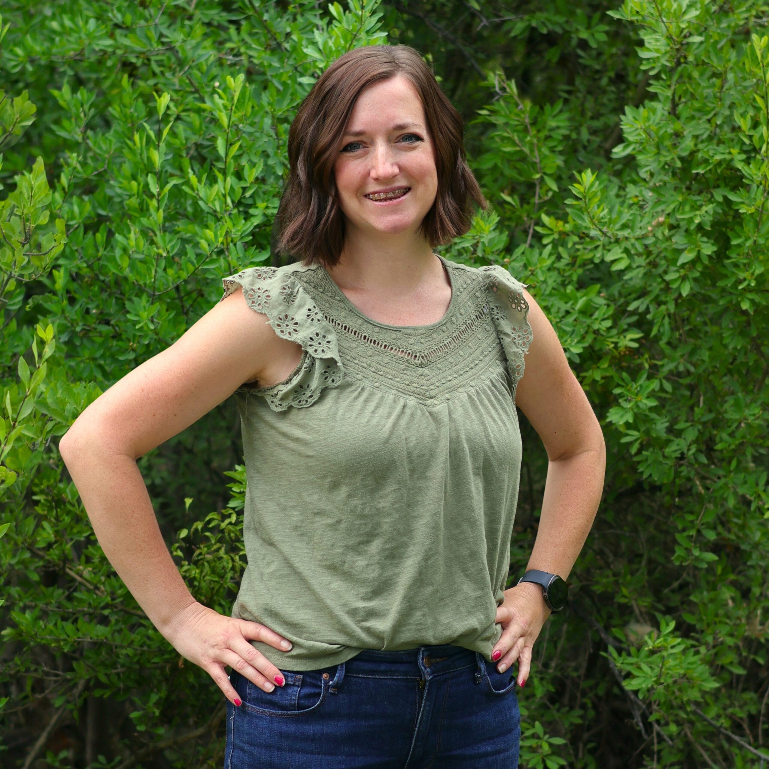A woman with short brown hair stands with her hands on her hips in front of green shrubs, wearing a light green sleeveless top and dark jeans.