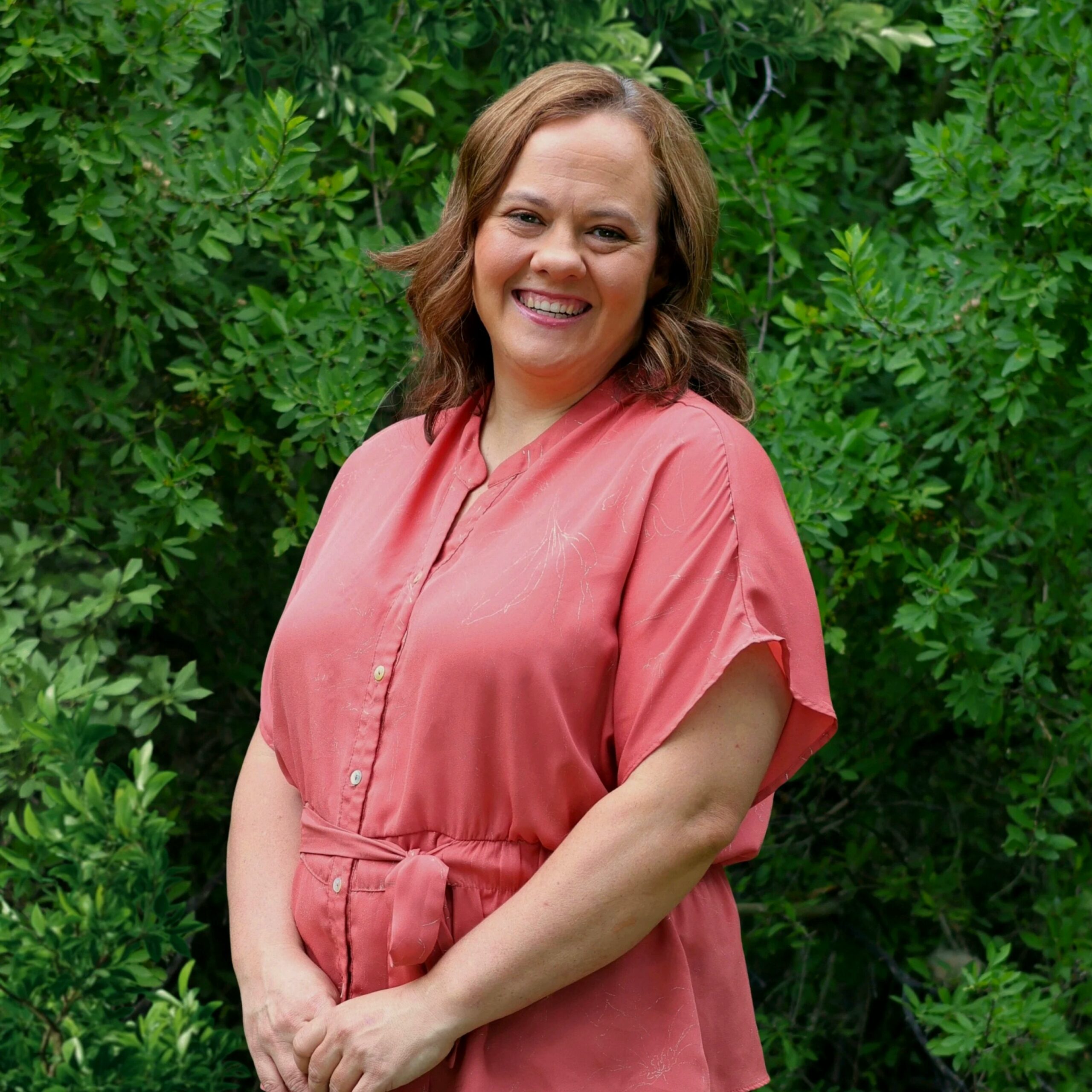 A smiling woman with long brown hair wears a coral button-up shirt tied at the waist, standing in front of dense green foliage.